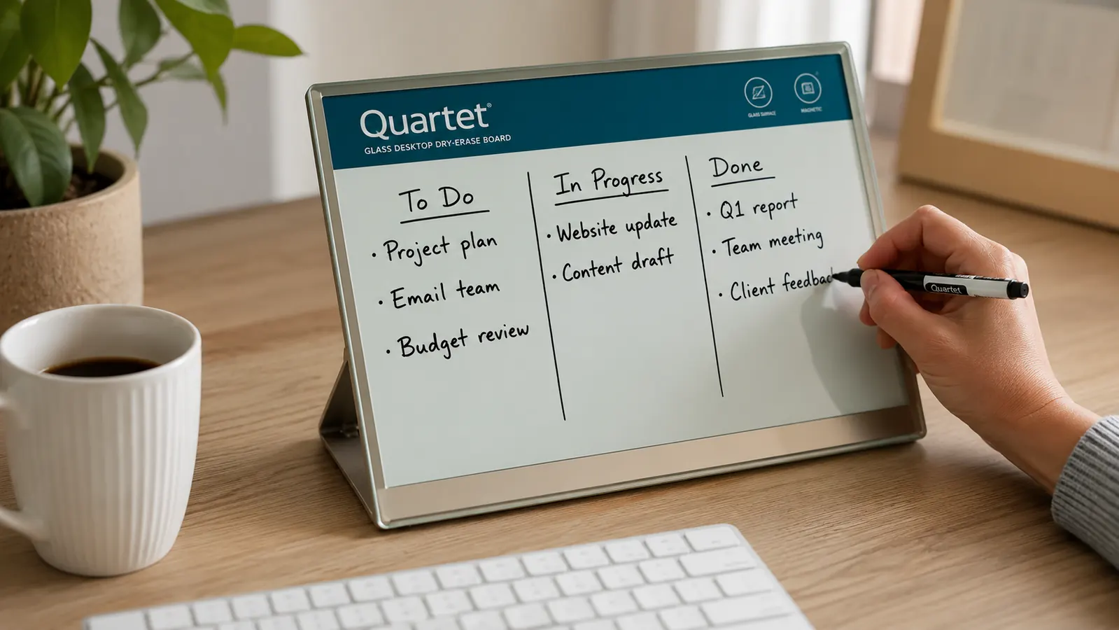 Hand writing the day's tasks on a glass desktop whiteboard propped against a monitor stand