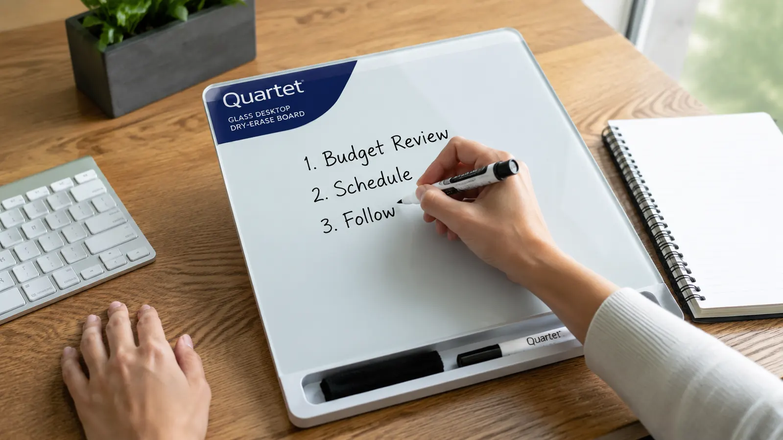 A hand writing a short numbered to-do list with a dry erase marker on a glass desktop whiteboard