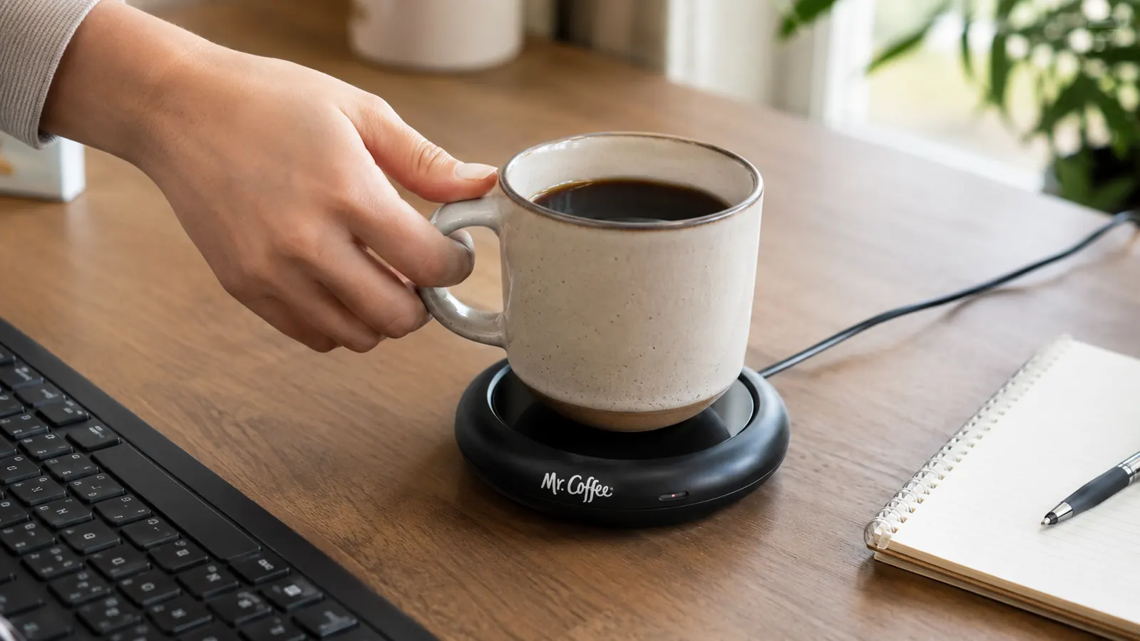 A hand placing a coffee mug onto an electric mug warmer on a desk