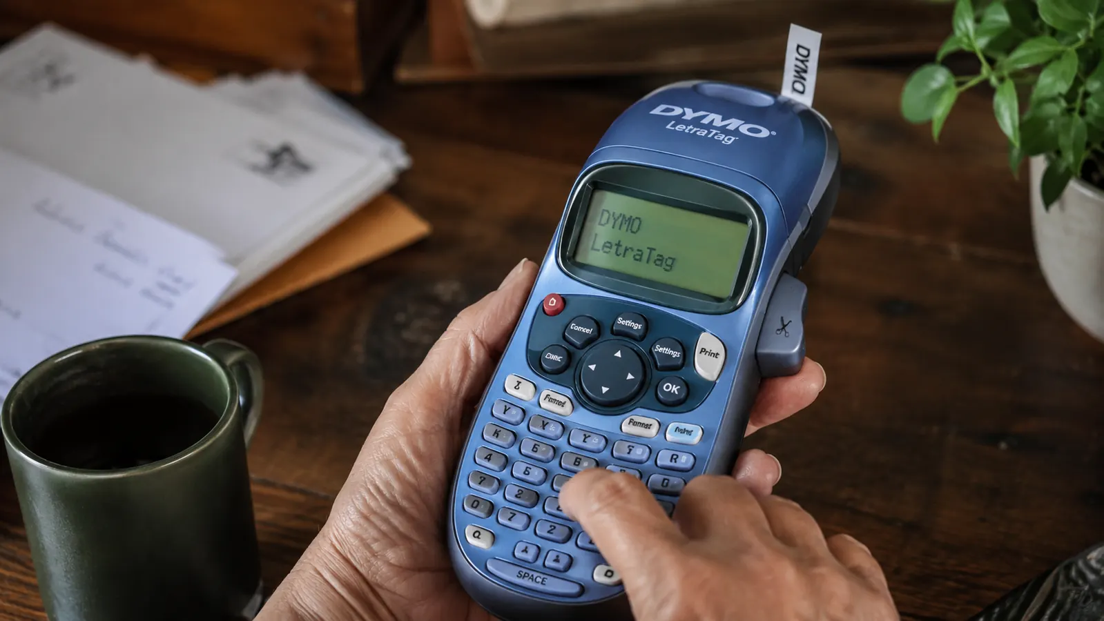 A hand pressing buttons on the DYMO label maker's alphabetical keyboard while printing a label