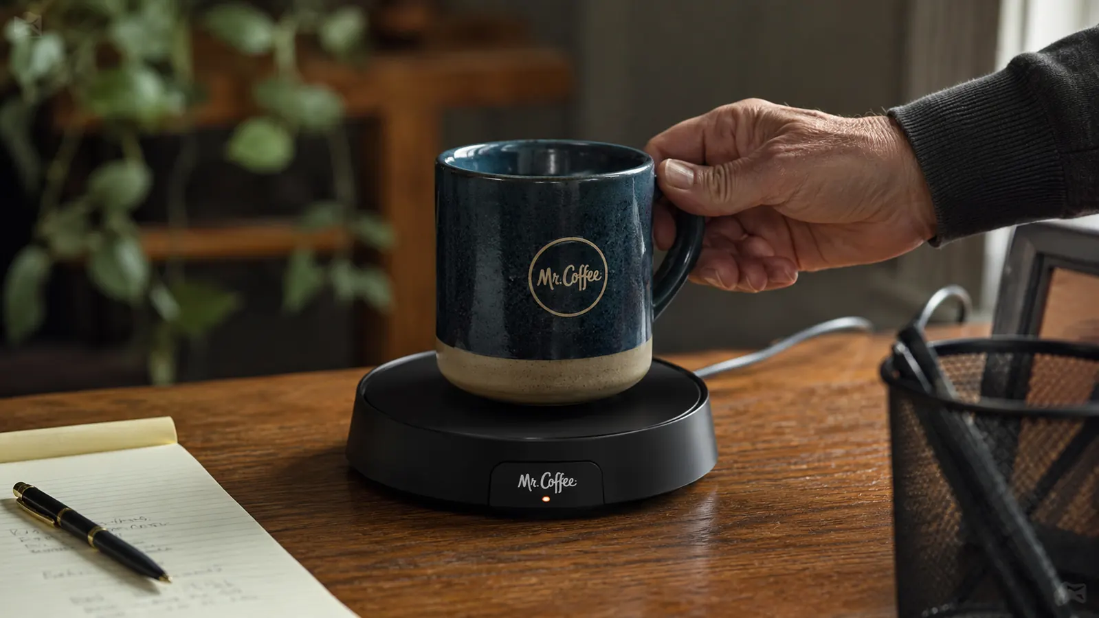Close-up of a hand setting a coffee mug onto the Mr. Coffee Mug Warmer's black round plate on a desk